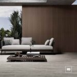 Modern outdoor seating area with a white cushioned sofa, gray pillows, and a low wooden coffee table on a slatted deck, backed by a tall brown slat wall and lush plants to the left.